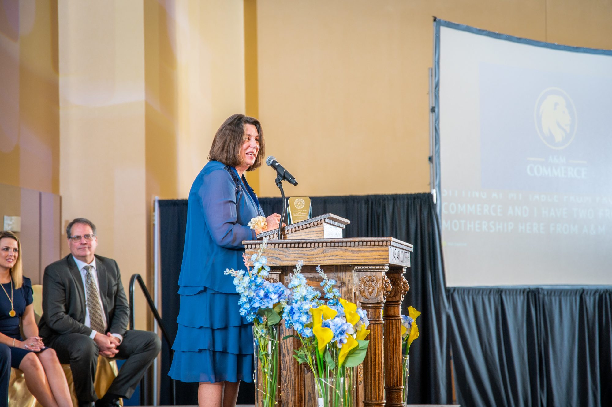 Awardee speaks to the crowd behind a podium, with two presenters sitting in chairs to her left.
