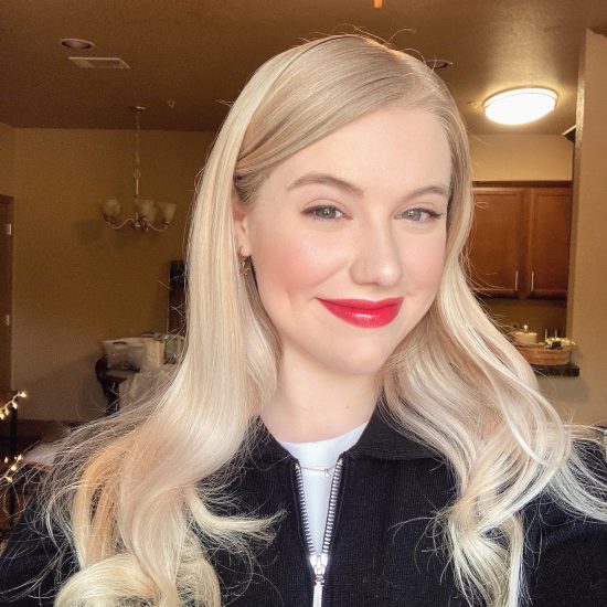 Portrait of a smiling woman with long, wavy platinum blonde hair, wearing a black zip-up top. She has bright red lipstick and is posing indoors, with soft lighting illuminating her face. The background includes part of a living space, with wooden cabinets and a light fixture visible.
