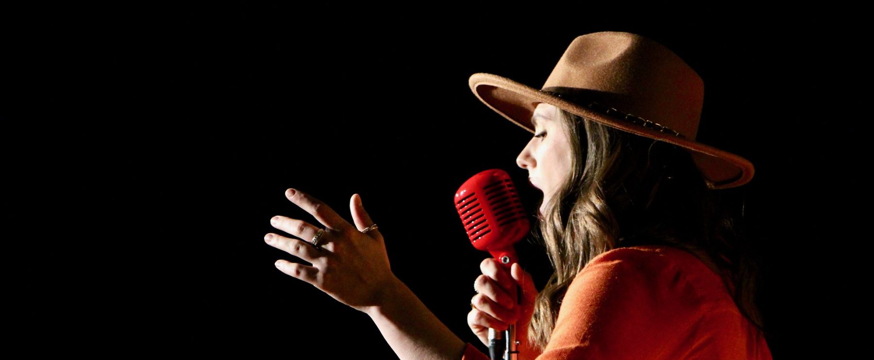 A person wearing a tan hat and an orange shirt holds a red vintage-style microphone and gestures with their hand while singing against a black background.
