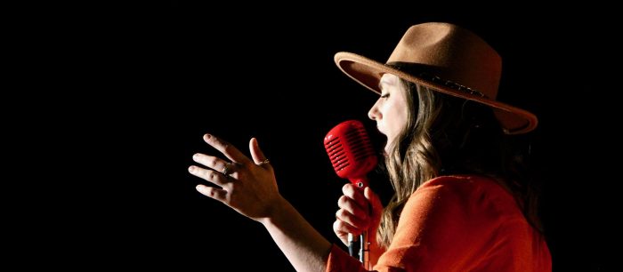 A person wearing a tan hat and an orange shirt holds a red vintage-style microphone and gestures with their hand while singing against a black background.