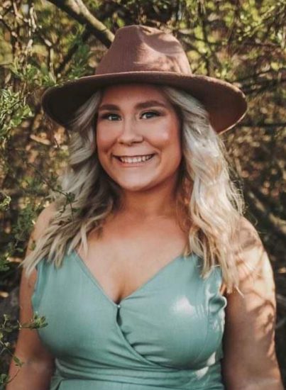 A woman with long blonde hair is smiling outdoors. She is wearing a wide-brimmed brown hat and a light blue sleeveless dress. The background features leafy trees and sunlight filtering through the branches.