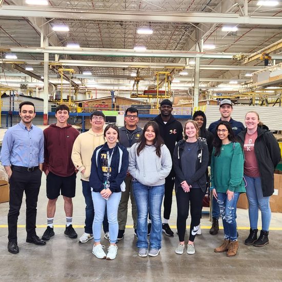 A group of twelve people stand in a warehouse, posing for a photo. They are dressed casually and smiling at the camera. Industrial equipment and shelving are visible in the background, under a high ceiling with exposed structural elements.