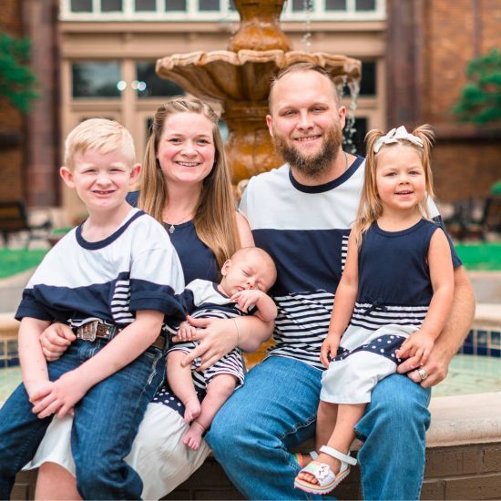 A family of five sits together on a low brick wall in front of a fountain. They are all smiling, and the parents are holding their three children. Everyone is dressed in coordinating navy blue and white outfits. The background features a brick building and greenery.