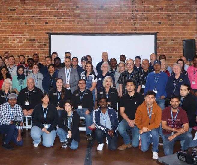 A large group of people posing for a group photo in front of a brick wall and a projector screen. Some are standing while others are sitting or kneeling in front. They are wearing casual and business-casual attire, with many wearing badges.
