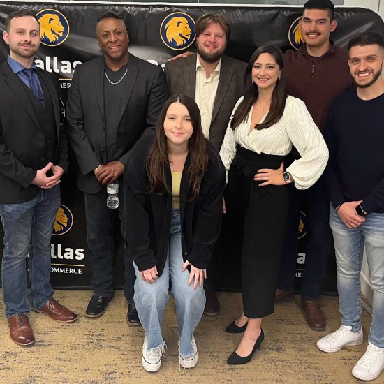 A group of seven people is posing for a photo in front of a backdrop with logos. Six individuals are standing, and one is crouching in the front. They are all smiling and dressed in casual to business casual attire.