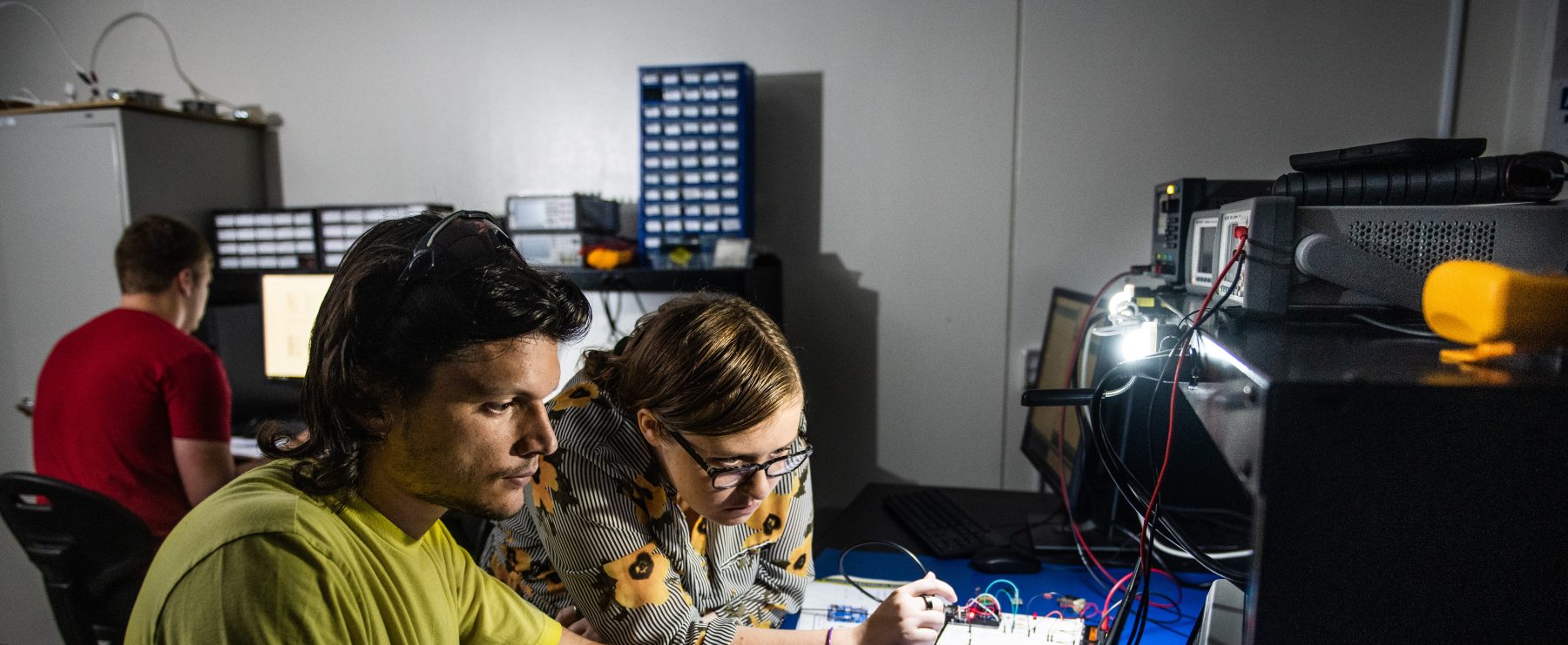 Two individuals are working together at a desk equipped with electronic components and a laptop. The man uses the laptop, while the woman interacts with electronic devices and wires while looking at the computer screen. Another person is in the background focusing on their computer. The room is dimly lit.
