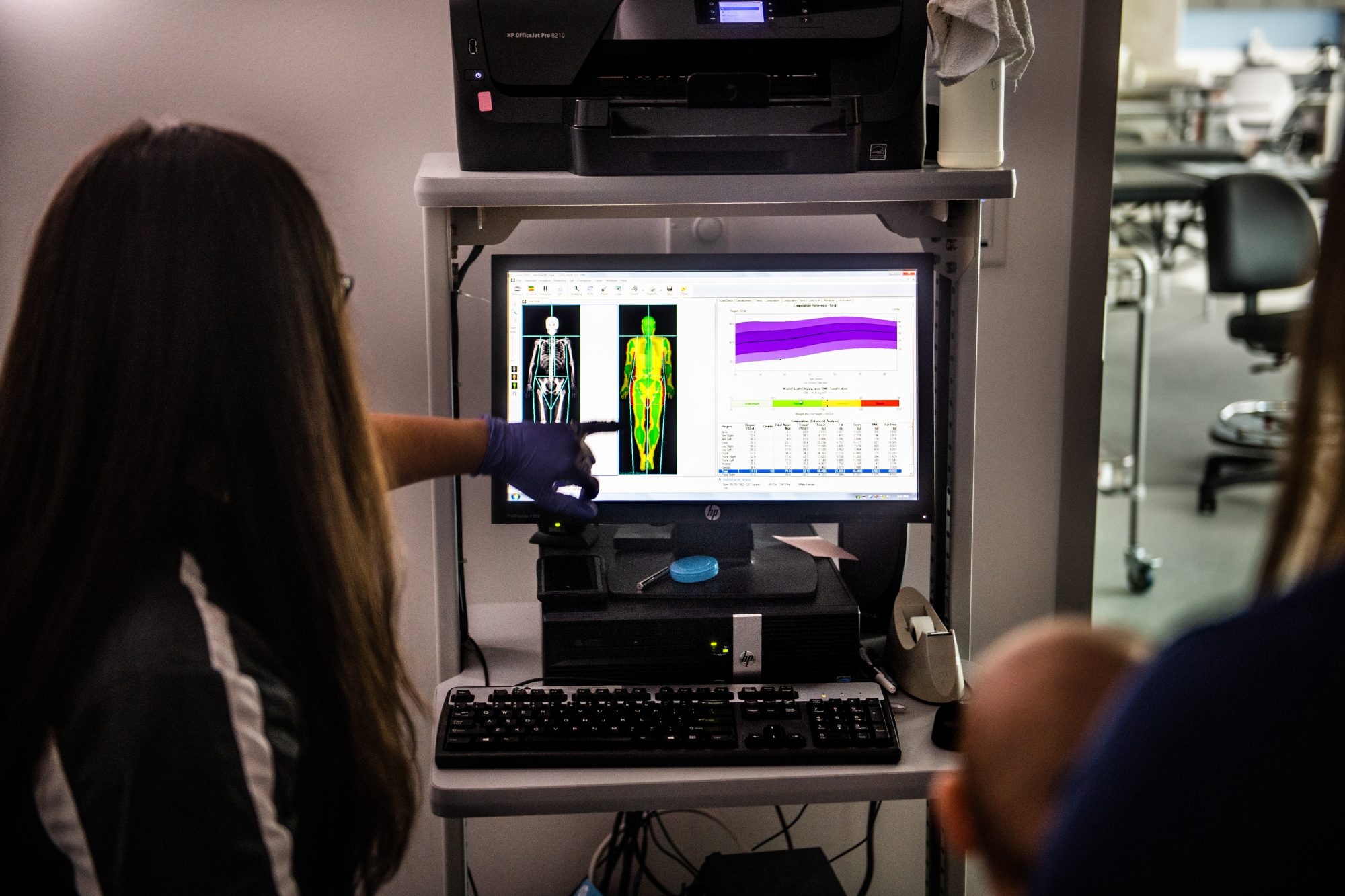 A researcher in gloves points at a computer screen displaying a full-body scan and data analysis, likely in a medical or scientific setting, with another person observing nearby.
