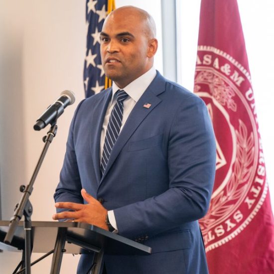 A man in a blue suit and striped tie speaks into a microphone at a podium. An American flag and a maroon flag with a university seal are visible in the background.