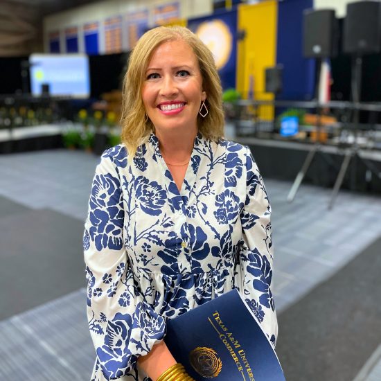 A woman with shoulder-length blonde hair wearing a white dress with blue floral patterns is holding a blue Texas A&M University diploma folder. She is standing in a large indoor venue with a stage and seating area in the background, and she is smiling at the camera.