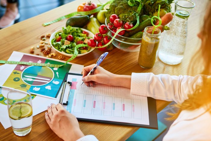 oman dietitian in medical uniform with tape measure working on a diet plan sitting with different healthy food ingredients in the green office on background. Weight loss and right nutrition concept