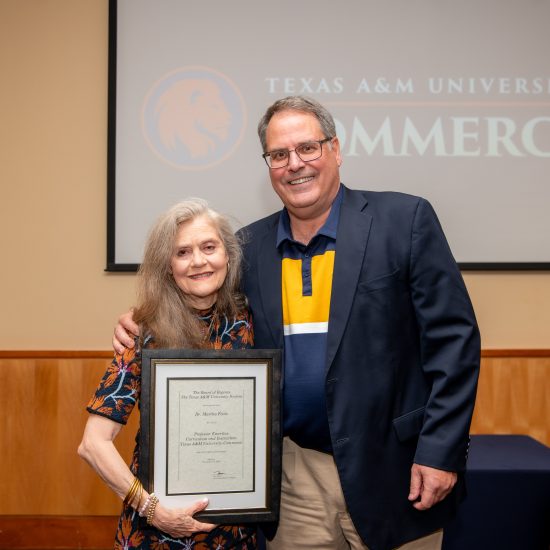 A man and a woman stand closely together, both smiling. The man is wearing a blazer over a collared shirt, and the woman is holding a framed certificate of recognition. Behind them is a projected A&M-Commerce logo.