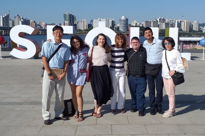 A group of seven people stands together, smiling towards the camera. Behind them, large white letters spell out "SEOUL". The backdrop features a river and a modern city skyline with numerous high-rise buildings under a clear blue sky.