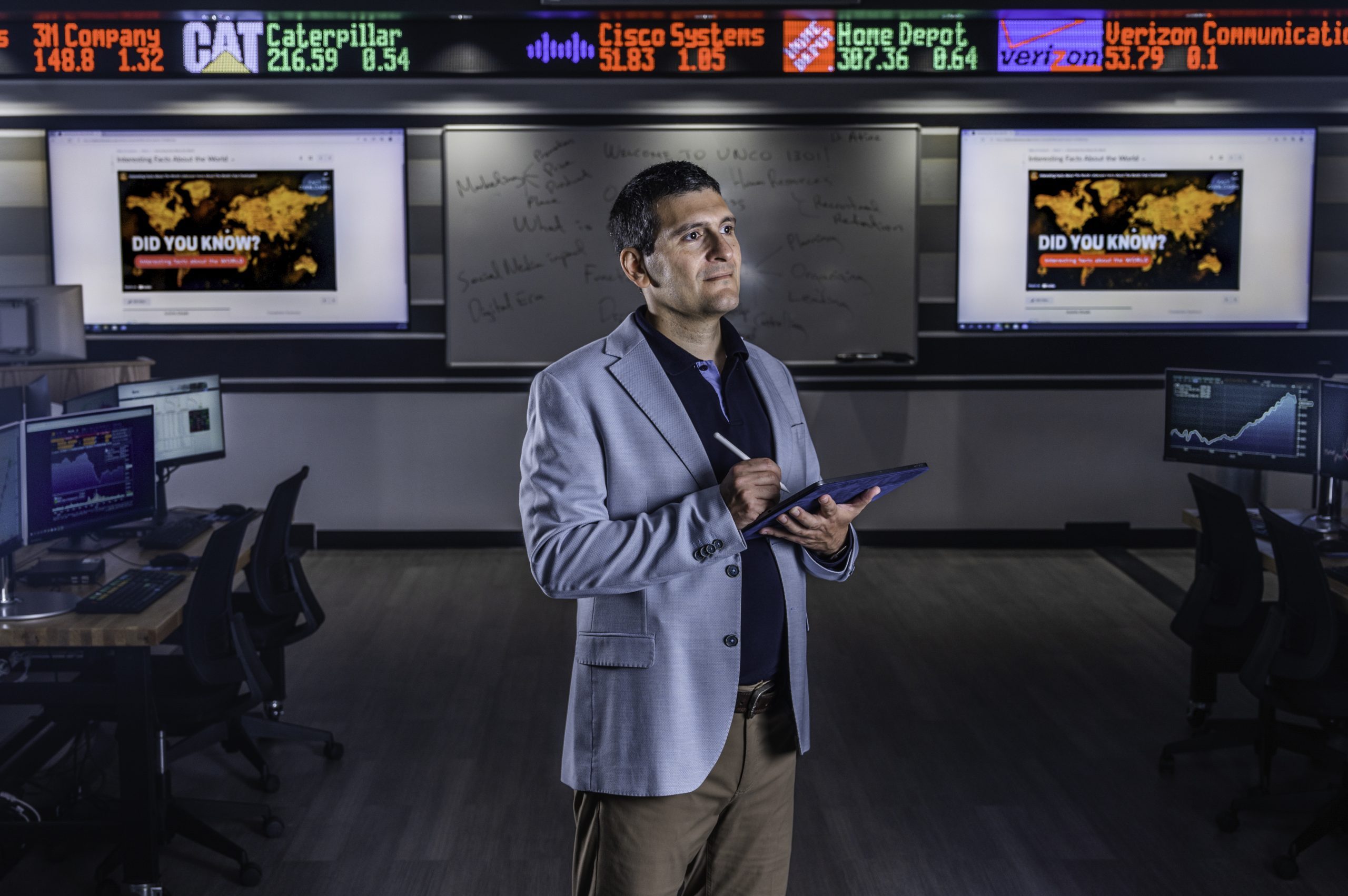 A man in a blazer stands in a modern office with multiple computer monitors and flat screens behind him displaying a world map and the text "Did You Know?". He holds a tablet and looks thoughtful. An electronic stock ticker with various company names and indices runs above.