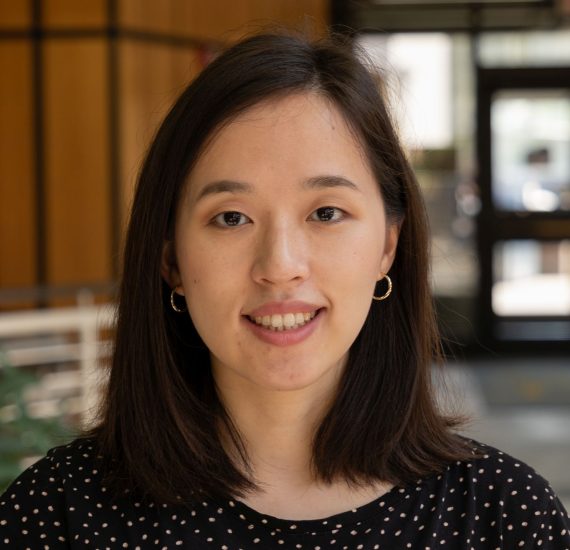 A young smiling female with dark brown shoulder length hair. She is wearing a black dotted shirt. In the background appears to be a door entrance and sits.