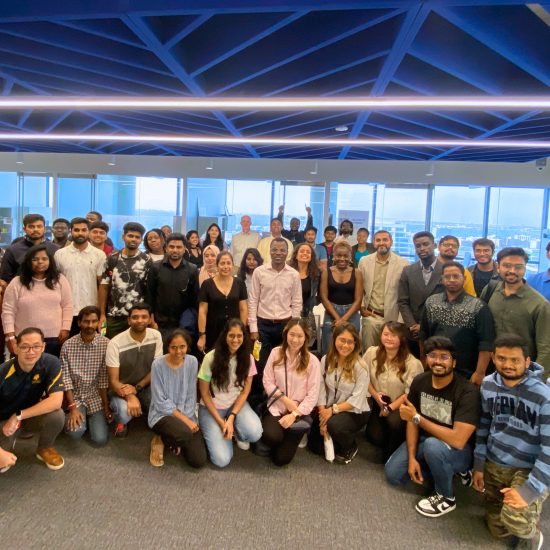 A large group of people posing together in an office setting with large windows in the background. The group consists of both men and women of various ages, standing and seated in multiple rows, smiling at the camera. The ceiling features a geometric blue design.