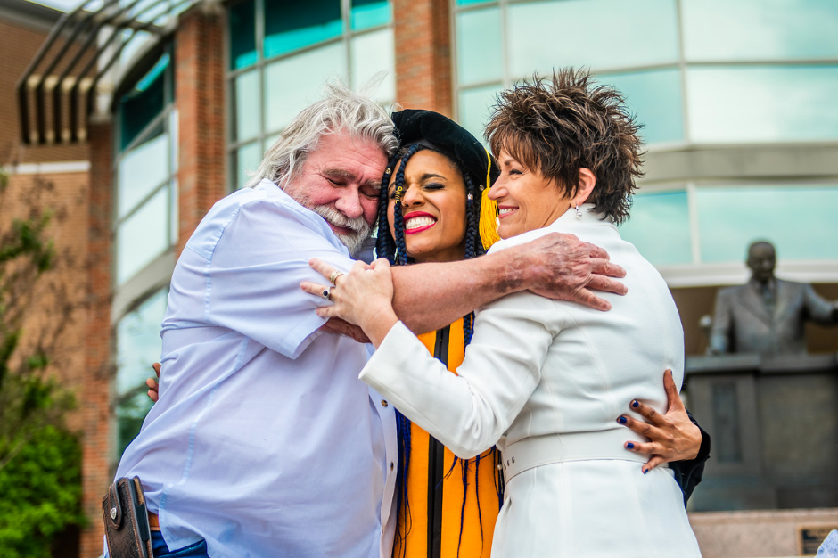 Three people are celebrating outdoors. A graduate in a yellow gown and black cap is embraced by an older man with white hair and a woman with short hair, both smiling. They stand in front of a modern building with large windows.