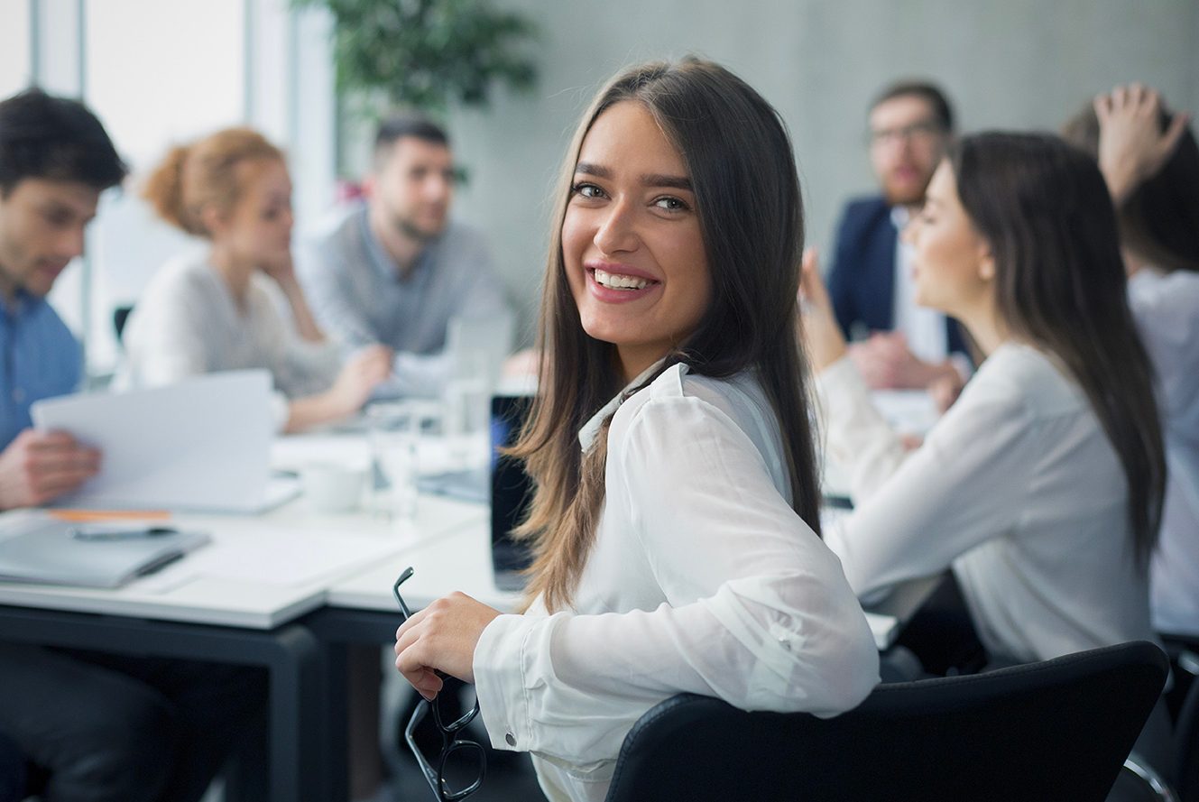 A woman with long brown hair and a white blouse is smiling at the camera in a modern office meeting room with several other people behind her.
