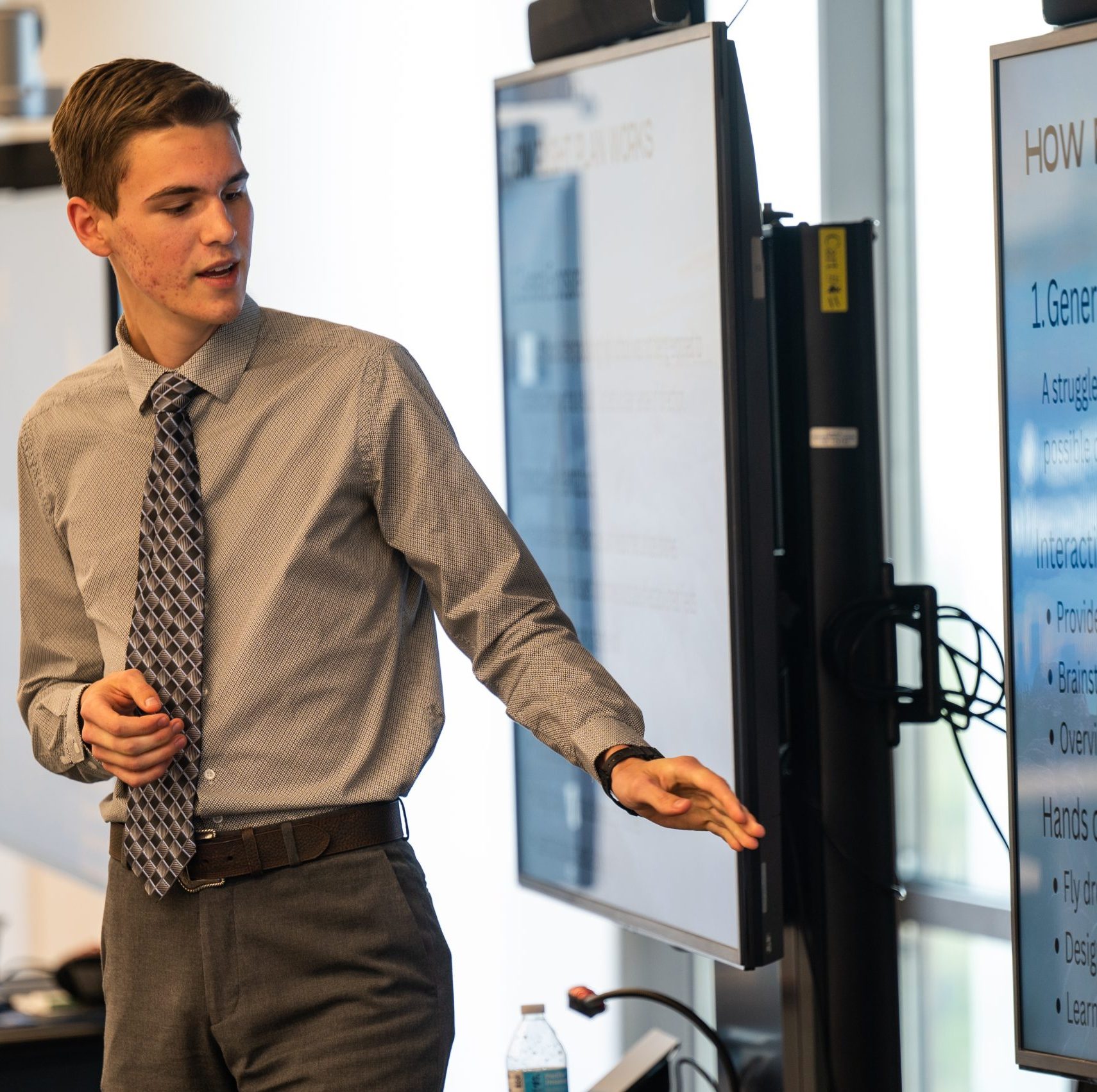 A young male in business attire is giving a presentation in front of several different tvs.