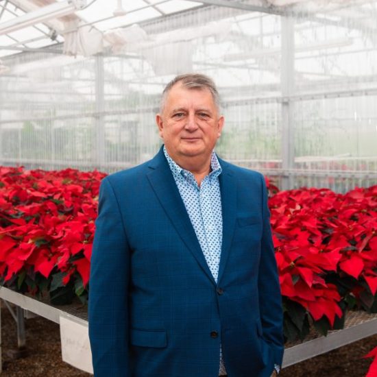 A man in a blue suit and polka-dotted shirt stands in a greenhouse filled with red poinsettia plants. He has short, gray hair and a neutral expression. The greenhouse structure and mist are visible in the background.