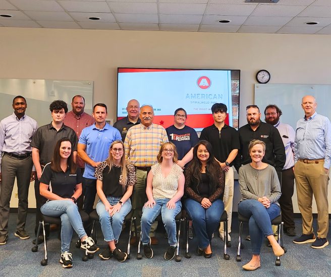 A group of fourteen people, some standing and some seated, pose for a photo in a room with a screen displaying "American Sprialweld Pipe." The background has whiteboards and a clock. The group includes both men and women of a wide range of ages and styles.