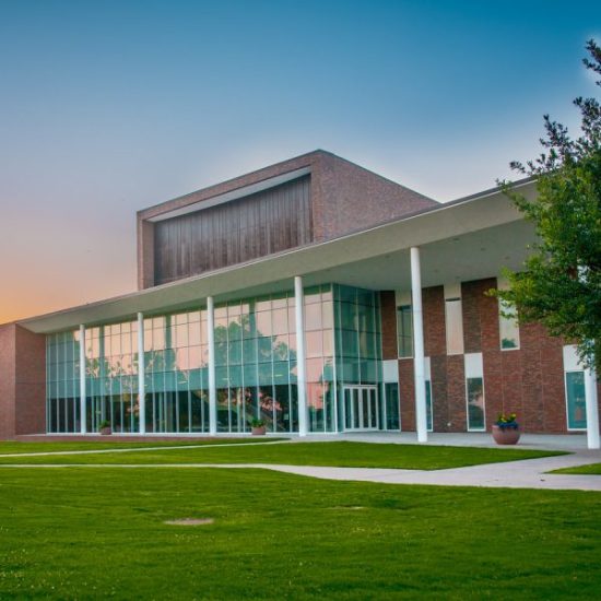 A modern building with large windows and brick accents, featuring an extended porch supported by white pillars with grass in front of the porch.