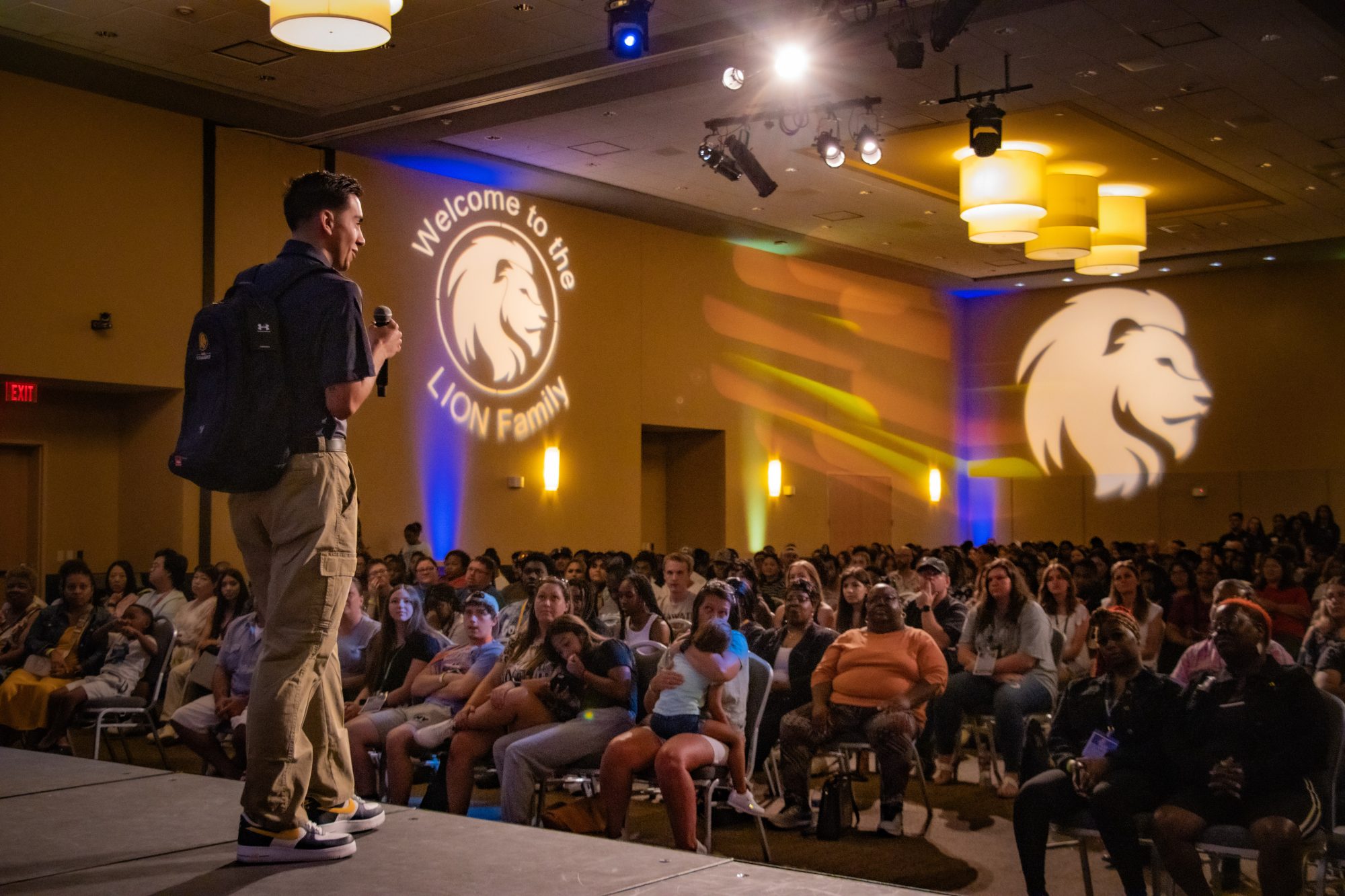 A student leading the ROAR presentation a stage with a large crowd in front of him. The ETAMU logo lit up in the background on the wall.