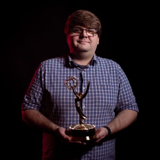 A young man stands against a dark background, holding his Emmy award confidently. The award's gold figure gleams, with one arm stretched upward and a globe encircled by wings at the top. The lighting highlights the person's face and the trophy.
