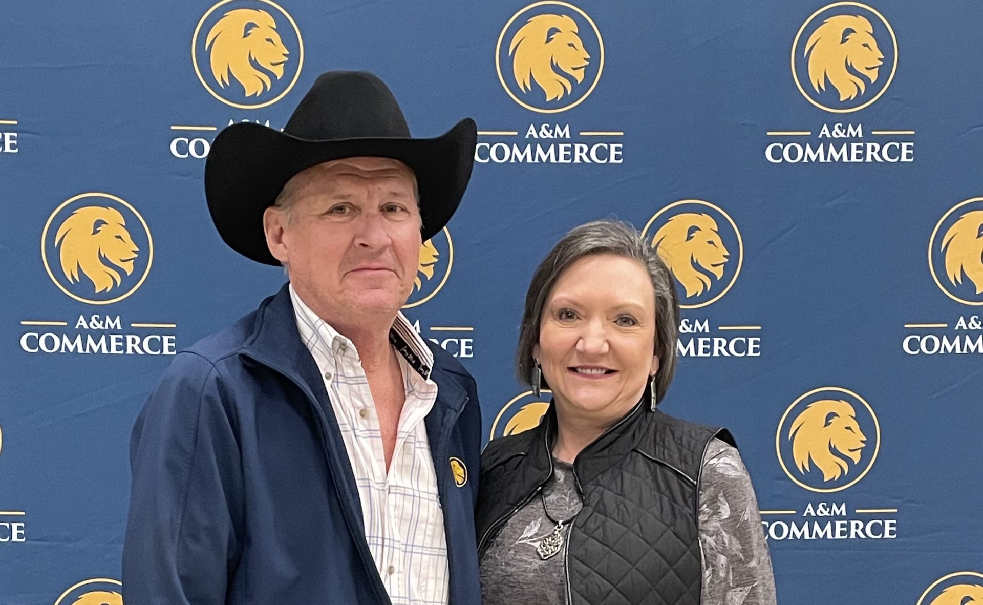 Scott and Margaret Stahl pose for a photo. Scott is wearing a black cowboy hat and the background is blue with several Lion head logos.
