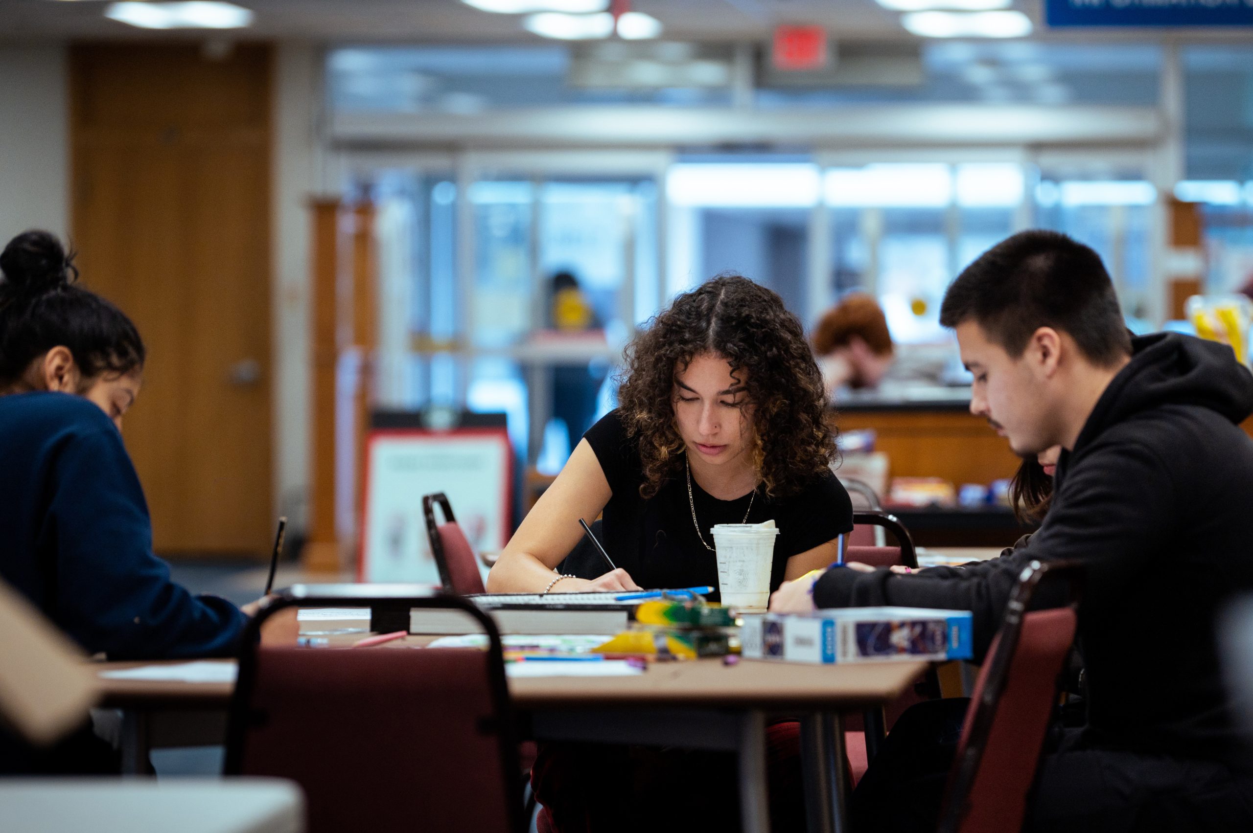 Three student sitting at a desk studying in what appears to be the library. The entrance in the background.