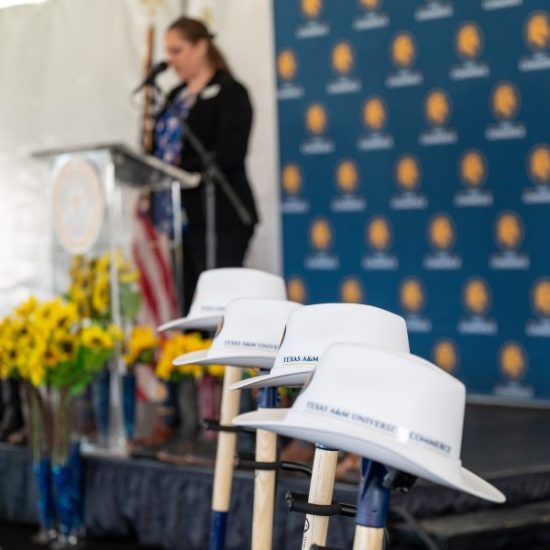 A woman speaks at a podium in the background, with an arrangement of sunflowers next to her. In the forefront, several ceremonial shovels with white hard hats labeled "Texas A&M University - Commerce" are arranged.