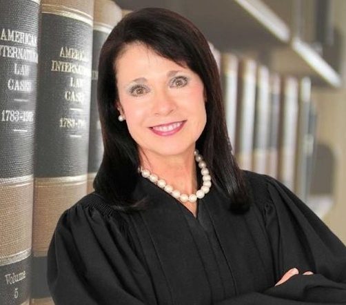 A person in a judge's robe leaning against bookshelves.