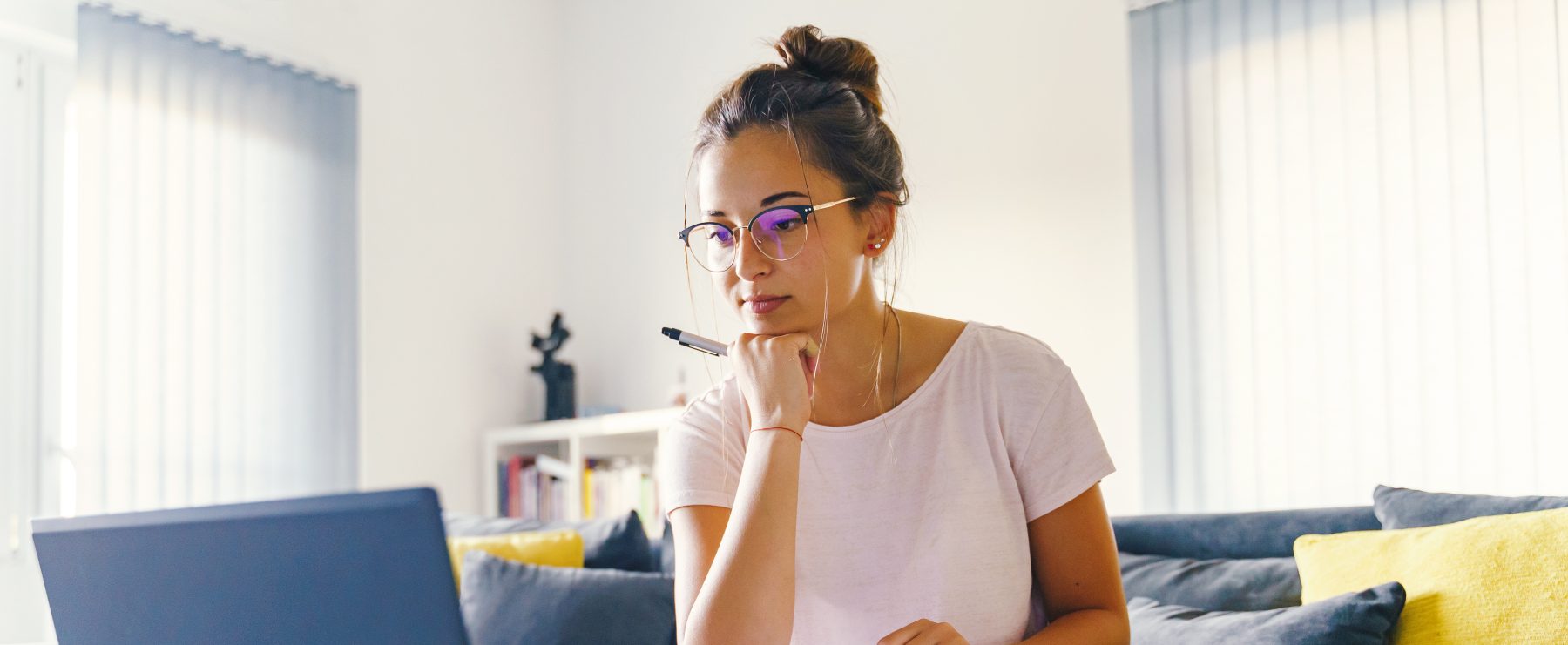 An adult learner sits on a couch in front of a laptop. Her chin is propped on her fist and she has a textbook and paper on her lap.