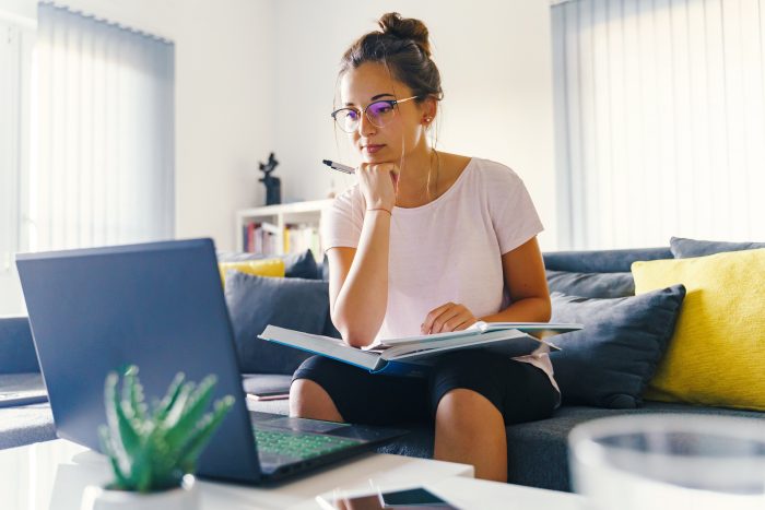 An adult learner sits on a couch in front of a laptop. Her chin is propped on her fist and she has a textbook and paper on her lap.