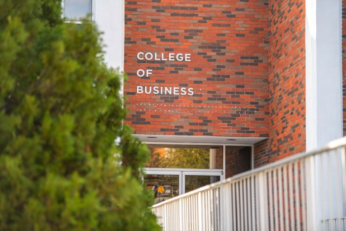 The entrance to a College of Business building, featuring a red and brown brick facade. Large white letters above the door read "College of Business." A forefront tree partially blocking the left side of the image, and a metal railing is visible on the right.