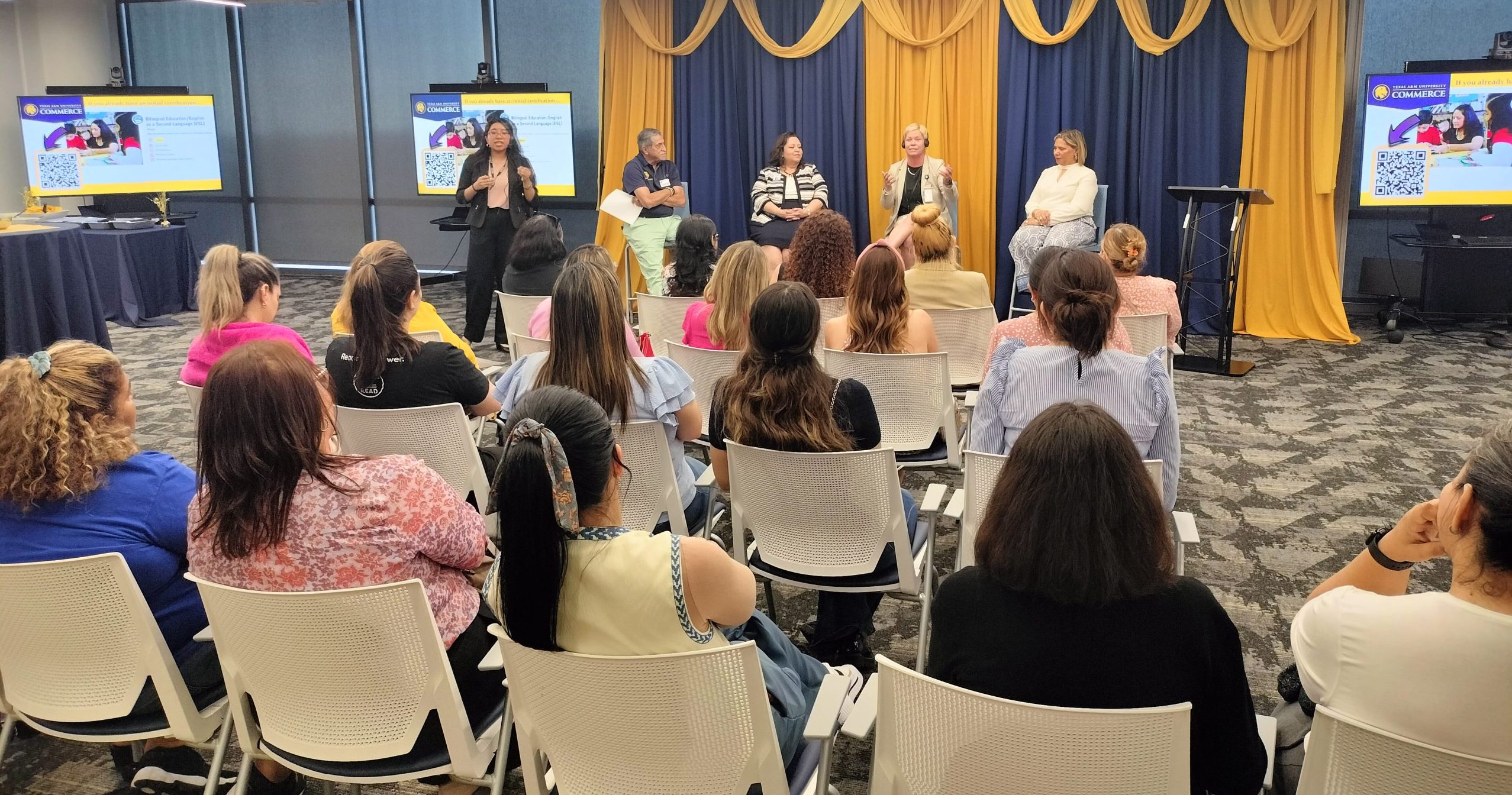 Audience faces a group of five panelists seated at the front of a conference room.