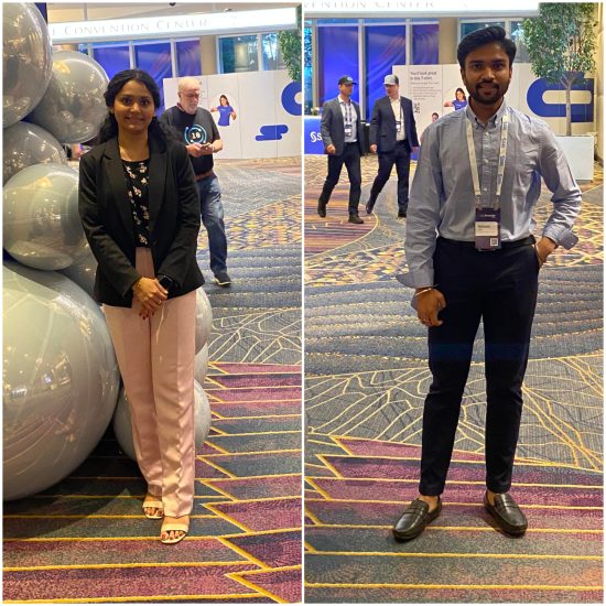 Two individuals at a convention center. On the left, a woman in a black blazer and pink pants stands in front of shiny silver spherical decorations. On the right, a man in a light blue shirt and dark pants stands on a patterned carpet. People walk in the background.