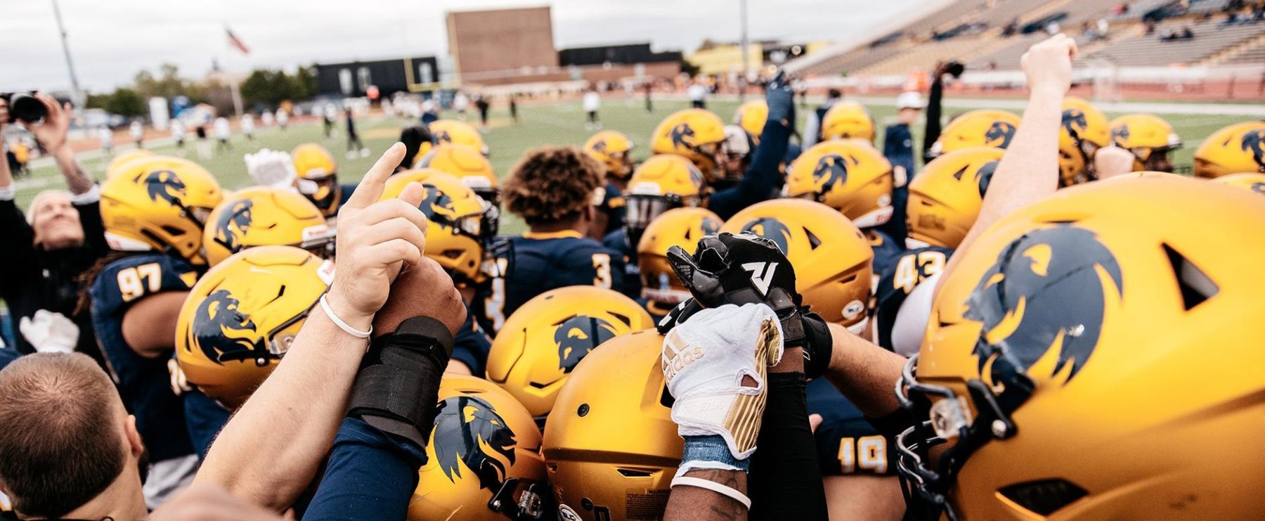 Football players group up before entering the field. Photo shows the tops of several players' helmets; some players have their arms raised. Football stadium is shown in the background.