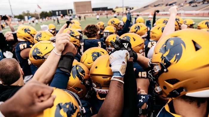 Football players group up before entering the field. Photo shows the tops of several players' helmets; some players have their arms raised. Football stadium is shown in the background.