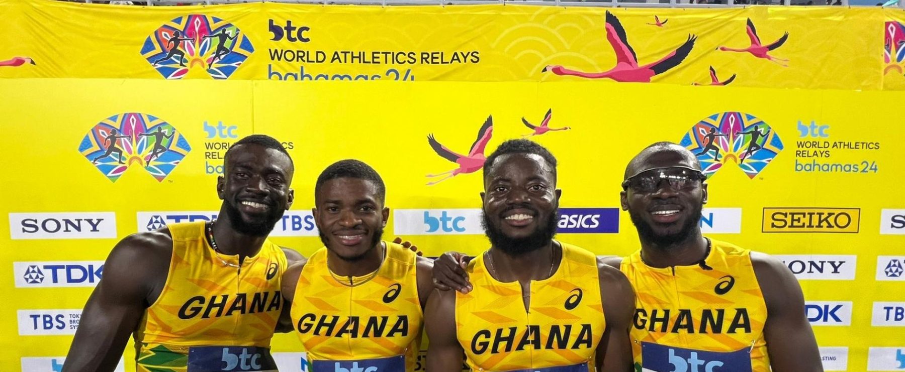 Four athletes pose before a yellow background, wearing their yellow Ghana jerseys and smiling with their arms around each other.