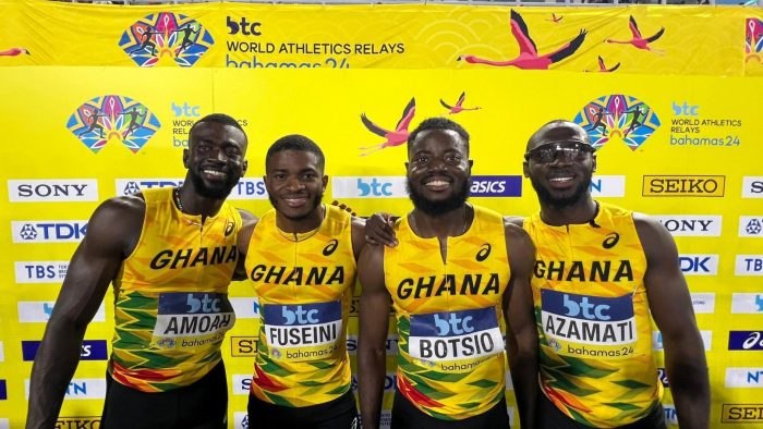 Four athletes pose before a yellow background, wearing their yellow Ghana jerseys and smiling with their arms around each other.