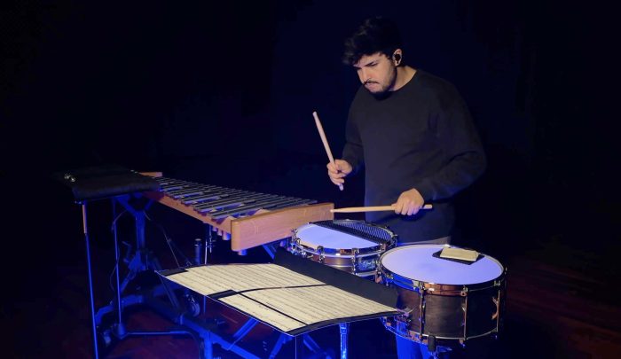 A musician dressed in black plays a marimba and two snare drums using drumsticks. Sheet music is spread out on a stand in front of him.