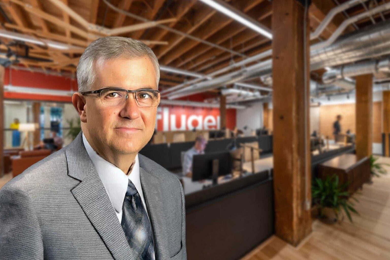 A man faces the viewer, with an office background behind him.