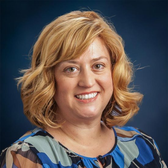 A smiling woman with shoulder length blonde hair, wearing a multicolor shirt, is set against a dark blue background.