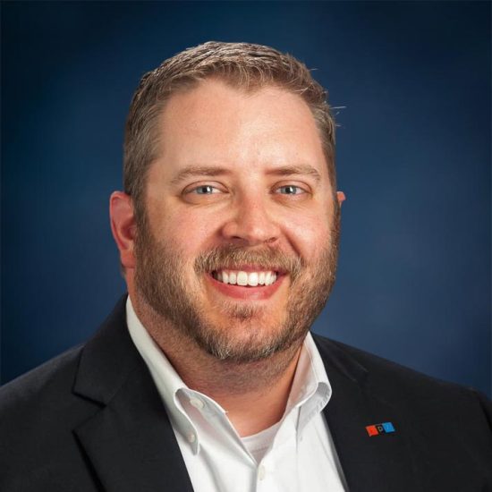 A smiling man with short light brown hair and a beard, wearing a white shirt and black blazer, is set against a dark blue background.