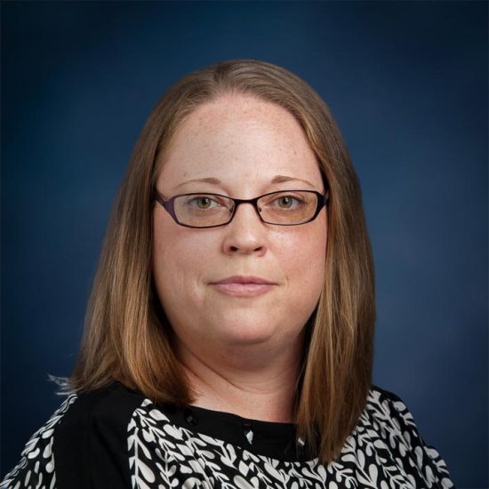 A woman with straight, shoulder-length light brown hair, wearing glasses and a black-and-white patterned blouse, is set against a dark blue background.