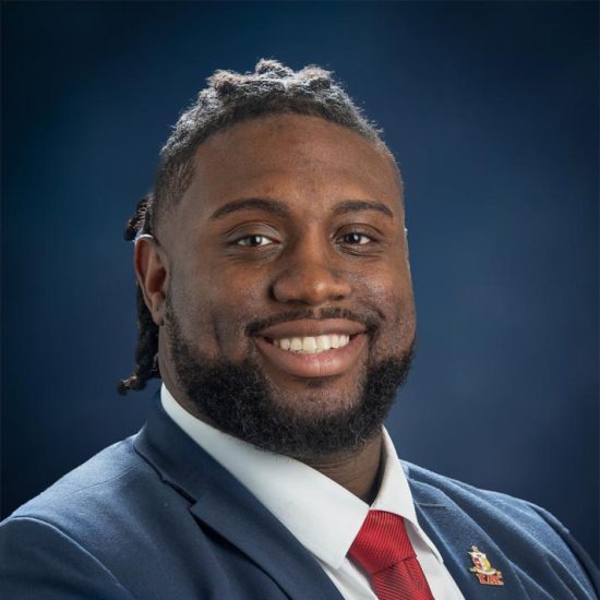 Portrait of a smiling man with a short hair and a beard, wearing a white shirt and blue blazer, is set against a dark blue background.
