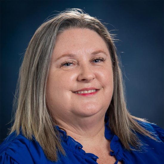 A smiling woman with shoulder length blonde straight hair, wearing a navy blue shirt, is set against a dark blue background.