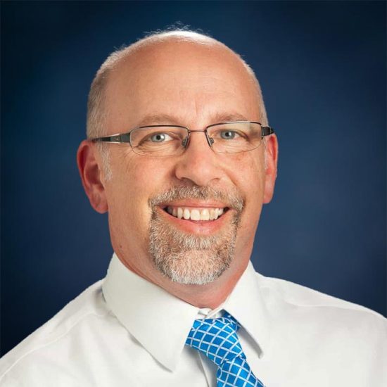 A man with glasses, wearing a white shirt and blue patterned tie, smiles widely in a formal portrait against a blue backdrop.