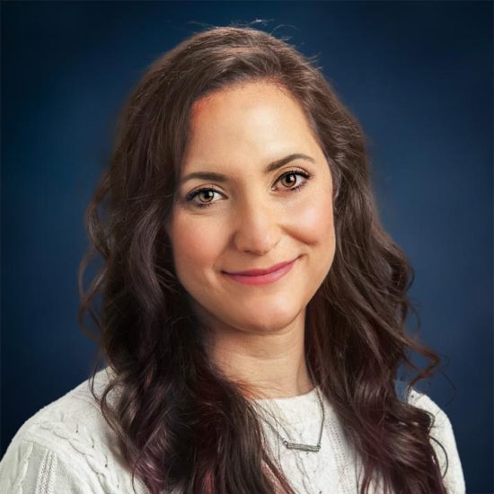 Professional portrait of a woman with brown hair, smiling and wearing a white sweater with a necklace, set against a dark blue background.