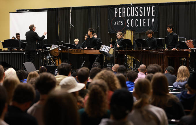 A group of musicians performs on stage with various percussive instruments, including a steel drum and marimbas, in front of an audience. A conductor in a black suit stands to the left, leading the ensemble. A "Percussive Arts Society" banner hangs in the background.